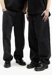 Jeans di denim neri a gamba larga con una finitura leggermente sbiadita, dotati di tasche laterali e dettagli sfrangiati, abbinati a scarpe stringate nere.
