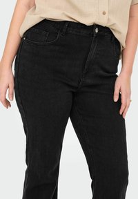 Los jeans de denim negro presentan un clásico diseño de cinco bolsillos con cremallera y cierre de botón. La tela tiene una textura suave y un ligero estiramiento.