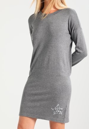 Kvinde iført en langærmet, grå strikket kjole med en sølvglitrende stjerne nær kanten på venstre side.