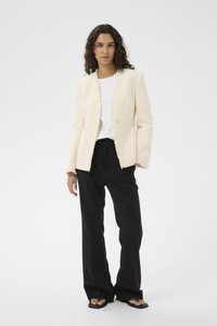 Blazer in cremiger Textur mit ausgefransten Kanten, Ein-Knopf-Verschluss, kombiniert mit schwarzen, weit geschnittenen Hosen und schwarzen flachen Sandalen.