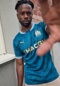 Le maillot de football bleu présente un design texturé, un logo doré et du texte. Des accents blancs sur les manches, avec un col arrondi et une coupe slim.
