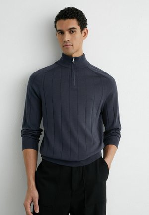 Tengerészkék negyed cipzáras pulóver bordázott textúrával és függőleges csíkokkal, magas gallérral és testhez simuló ujjal, fekete nadrággal párosítva.