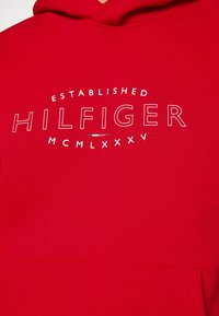 Sweat à capuche rouge en tissu doux présentant le mot "HILFIGER" en blanc, avec "ESTABLISHED" et "MCMLXXXV" disposés en cercle au-dessus.