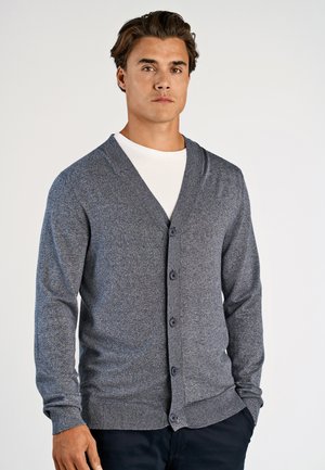Homme debout, les mains dans les poches, portant un cardigan gris boutonné par-dessus une chemise blanche sur un fond uni.
