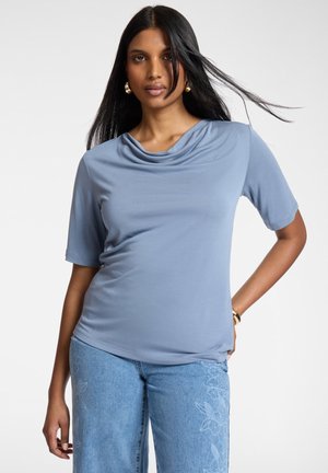 Donna con capelli lunghi neri che indossa un top azzurro chiaro con collo a scialle e jeans azzurri chiari ricamati, posando con una mano sui fianchi.