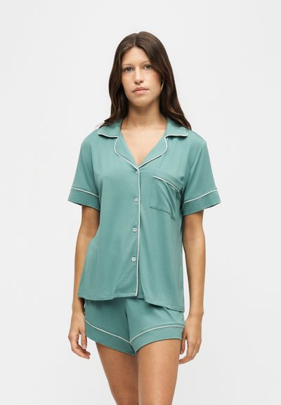 Femme portant une chemise bleu sarcelle à manches courtes avec passepoil blanc et un short assorti sur fond blanc uni.