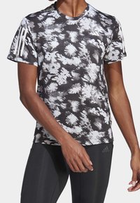 Camiseta atlética de manga corta con un patrón floral en blanco y negro, hecha de tejido texturizado. Presenta tres franjas blancas en el hombro izquierdo.