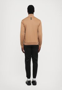 Vīrietis ģērbts kafejāda krāsas zip-up jakā ar melnām svītrām aizmugurē, melnās biksēs, baltās zeķēs un melnās sporta kurpēs, stāv ar seju prom.