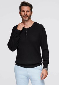 Maglietta nera a coste con maniche lunghe, scollo rotondo e pattina dei bottoni. Il modello indossa pantaloni azzurri chiari e un orologio al polso sinistro.