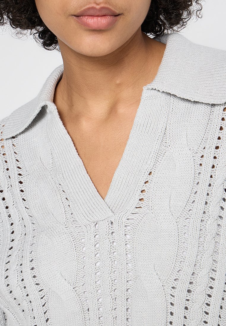 Donna che indossa un maglione grigio chiaro a trecce con un ampio colletto e scollo a V, che mostra dettagli del collo e della parte inferiore del viso.