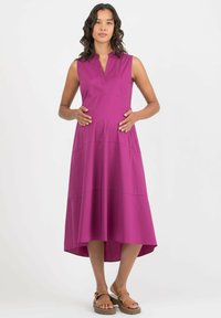 Ärmelloses, hochgeschlossenes lila Kleid mit ausgestellt geschnittenem Saum, aus weichem Stoff gefertigt. Das Model steht mit den Händen auf einem runden Bauch und trägt beiges Sandalen.