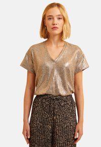 T-shirt en V métallique doré avec des manches courtes, présentant une texture côtelée et une finition brillante, associé à une jupe noire à motifs.