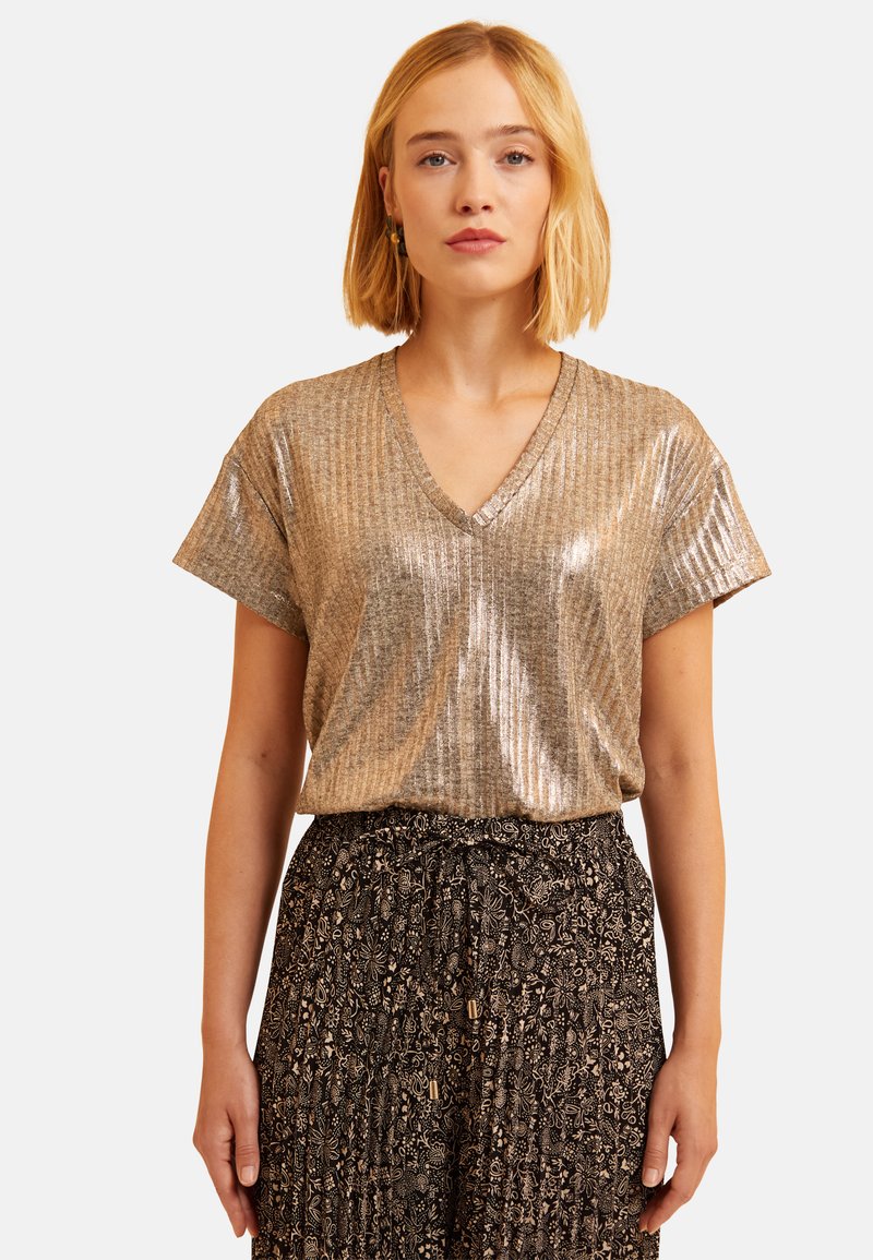 T-shirt en V métallique doré avec des manches courtes, présentant une texture côtelée et une finition brillante, associé à une jupe noire à motifs.