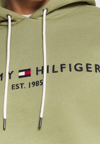 Kapuzensweatshirt aus olivgrünem Stoff, mit einem navyblauen gestickten Logo und weißen Kordeln mit silbernen Spitzen.