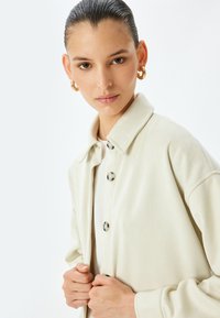 Giacca texturizzata beige chiaro con colletto e grandi bottoni, caratterizzata da una vestibilità ampia e maniche lunghe.