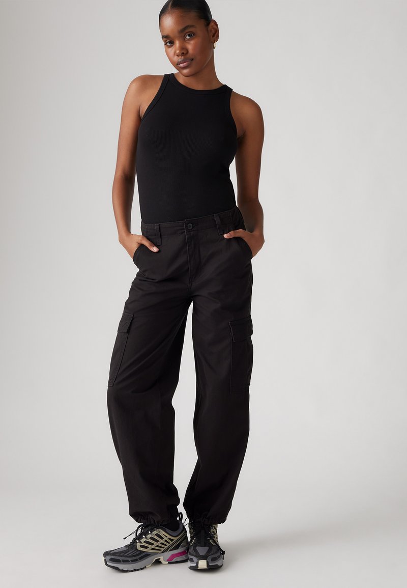 Débardeur noir à côtes associé à un pantalon cargo noir ample avec des poches latérales. Des baskets avec des accents gris et rose complètent le look.