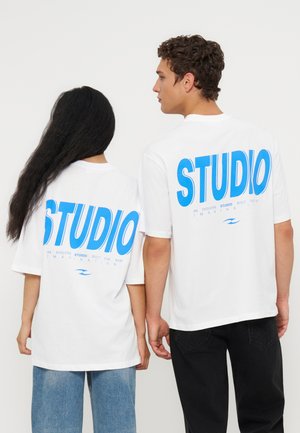 To personer iført hvide t-shirts med stor blå "STUDIO" tekst på ryggen, en i blå jeans og den anden i sorte bukser, vendt væk.