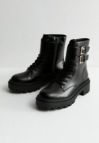Botas tobilleras de cuero negro con suela gruesa, parte delantera con cordones, cremallera lateral y dos hebillas en el puño. Textura suave y diseño elegante.