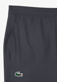 Tumši pelēki sporta šorti ar elastīgu jostasvietu, sastāv no smalkas tekstūras raksta un attēlo zaļu un baltu krokodila logo kreisajā kājā.