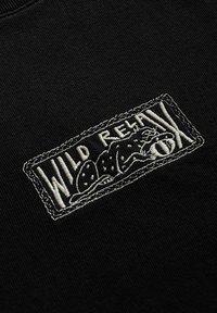 Czarna tkanina o teksturowanej powierzchni z białą haftowaną naszywką. Naszywka z napisem "WILD RELAX" i stylizowanym wzorem lisa.