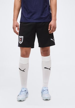 Άνδρας με μαύρα ποδοσφαιρικά σορτς με το έμβλημα της Αυστρίας, λευκές κάλτσες Puma μέχρι το γόνατο, ανοιχτό μπλε παπούτσια ποδοσφαίρου και σκουρό μπλε μπλουζάκι.