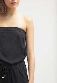 Combinaison noire sans bretelles avec un encolure froncée, une taille à cordon de serrage et une texture douce et lisse. Accentuée par des détails matériels subtils.