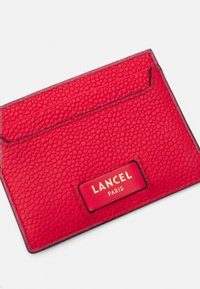 Portafoglio in pelle rossa con finitura a granulato, dotato di etichetta con logo dorato. Include più slot per carte e una forma rettangolare compatta.