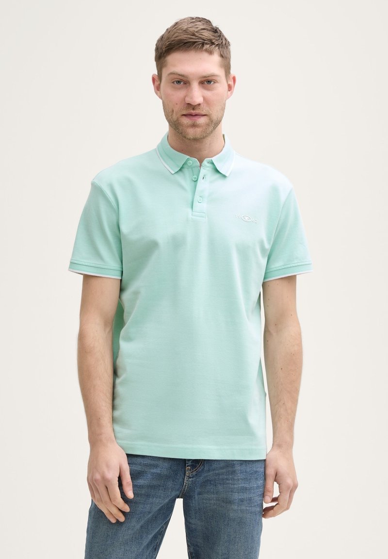 Mintgrünes Polohemd mit Knopfleiste, kurzen Ärmeln und kontrastierendem Kragenbesatz. Getragen mit blauen Jeans, die ein gesticktes Logo aufweisen.