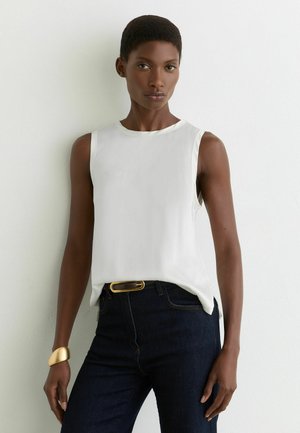 Kobieta w białej, bezrękawnikowej bluzce i ciemnych dżinsach, stojąca na tle jednolitej białej ściany, z bransoletką i paskiem w złotych detalach.