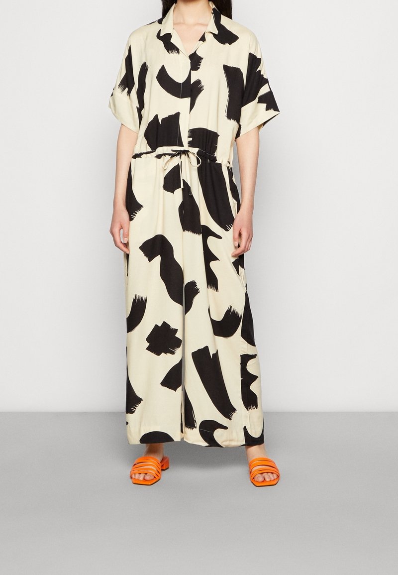 Robe maxi en tissu crème avec de grands motifs en traits de pinceau noirs, dotée d'un col, de manches courtes et d'une taille cintrée avec un cordon de serrage. Sandales plates orange.