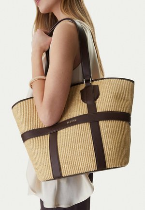 Donna che porta una grande borsa a tracolla beige intrecciata con manici e rifiniture in pelle marrone scuro sulla spalla.