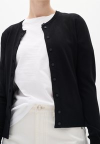 Person som bär en svart knäppt cardigan över en vit t-shirt som är stoppad i off-white högmidjade byxor mot en neutral bakgrund.