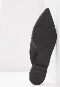 Chaussure plate noire pointue avec texture tissée, semelle montrant des motifs de chaussures de ballet en relief et le texte "PrettyBallerinas" visible en dessous.