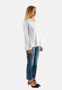 Blusa blanca de manga larga con cuello y botón en la parte frontal, combinada con vaqueros azules y tacones negros de punta. Tejido suave y corte relajado.