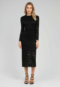Robe en dentelle noire à manches longues et à ourlet mi-genou. Présente un motif floral et des bords festonnés. Associée à des talons compensés noirs.