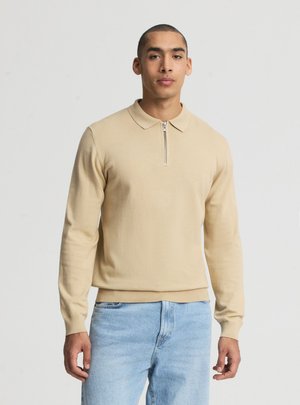 Uomo con maglione beige a manica lunga con zip e jeans azzurri, in piedi su uno sfondo chiaro e uniforme.