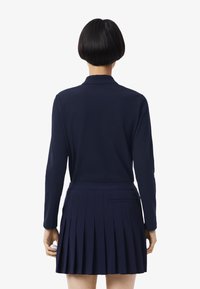 Marineblaues Langarmpolo-Shirt, getragen mit einem plissierten Rock, mit Kragen und einer taillierten Taille, das eine glatte Textur und ein maßgeschneidertes Design aufweist.