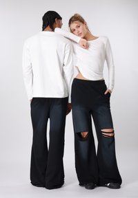 Λευκό μακρυμάνικο τοπ με καμπυλωτή ραφή στο τελείωμα, σε συνδυασμό με μαύρο παντελόνι με φαρδιά πόδια που έχει δύο μεγάλες κοψίματα στα γόνατα και τσέπες, και τα δύο από απαλό ύφασμα.