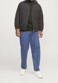 Czarna kurtka typu bomber na zamek, pod spodem zielony dzianinowy sweter, luźne niebieskie dżinsy i białe tenisówki. Gładkie tło.