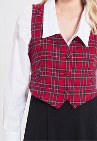 Czerwona kamizelka w kratę z ostrym dołem, z białą koszulą z kołnierzykiem pod spodem. Kamizelka ma dwa guziki i boczne kieszenie. Czarna spódnica poniżej.