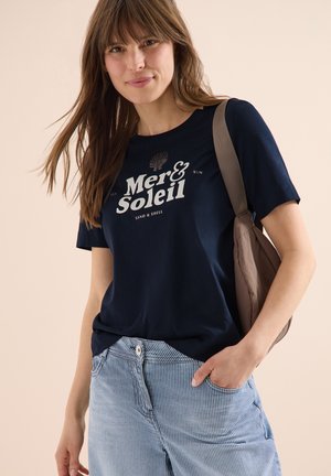 Vrouw met lang bruin haar die een marineblauw 'Mer & Soleil' T-shirt draagt, gestreepte blauwe broek en een beige schoudertas, met een lichte glimlach.