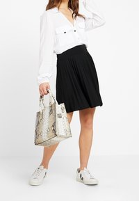 Väskan är mönstrad med ormskinn i beige och vitt, har en kompakt form med dubbla handtag. Modellen bär en vit skjorta, svart plisserad kjol och vita sneakers.