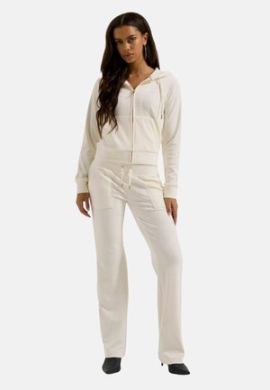 Femme aux longs cheveux foncés portant un sweat à capuche crème zippé et un pantalon large assorti avec poches avant et des chaussures noires à bout pointu.