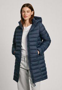 Manteau en duvet marine avec capuche, présentant des segments matelassés, une fermeture éclair sur le devant et des poches latérales. Porté sur une chemise blanche et un pantalon gris clair.