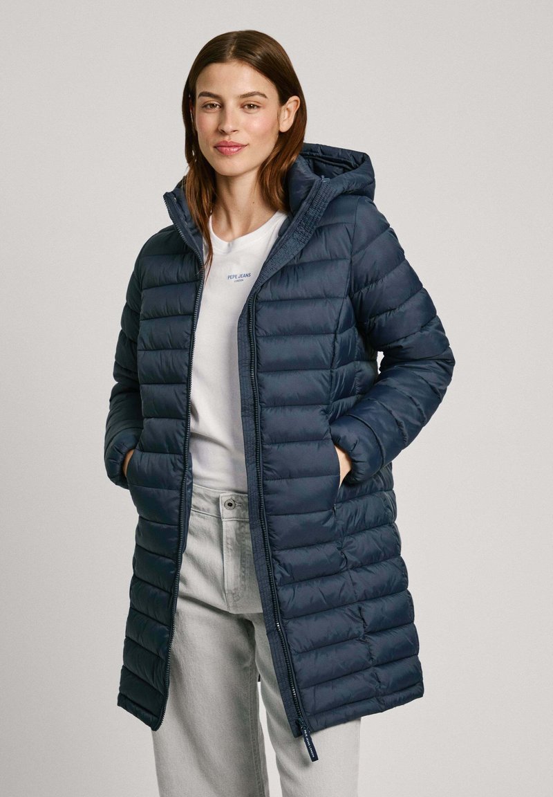Manteau en duvet marine avec capuche, présentant des segments matelassés, une fermeture éclair sur le devant et des poches latérales. Porté sur une chemise blanche et un pantalon gris clair.