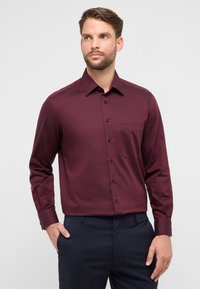 Bordeauxrode blouse met lange mouwen en een gestructureerd patroon, knoopsluiting aan de kraag, borstzak aan de linkerkant en donkere knopen. Gecombineerd met donkere broek.