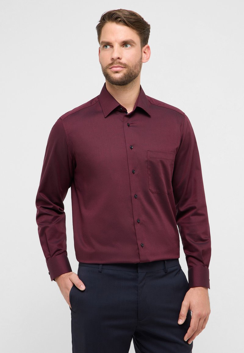 Bordeauxrode blouse met lange mouwen en een gestructureerd patroon, knoopsluiting aan de kraag, borstzak aan de linkerkant en donkere knopen. Gecombineerd met donkere broek.