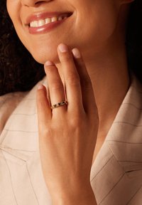 Gouden ring met vijf donkere stenen, gedragen op een bruine hand. De achtergrond toont een lichtgekleurde stof met subtiele strepen.