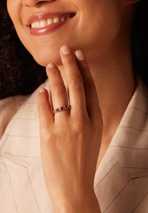 Goldring mit fünf dunklen Steinen, der an einer braunen Hand getragen wird. Der Hintergrund zeigt einen hellen Stoff mit feinen Streifen.