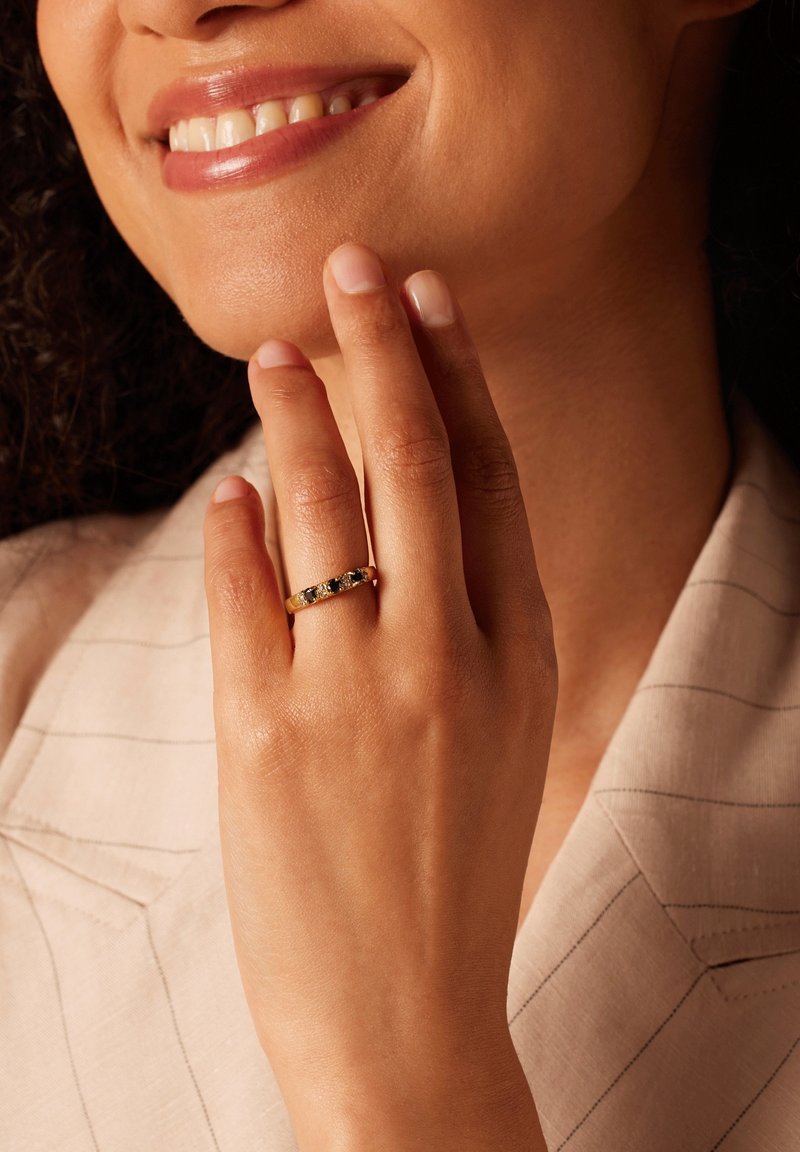 Gouden ring met vijf donkere stenen, gedragen op een bruine hand. De achtergrond toont een lichtgekleurde stof met subtiele strepen.